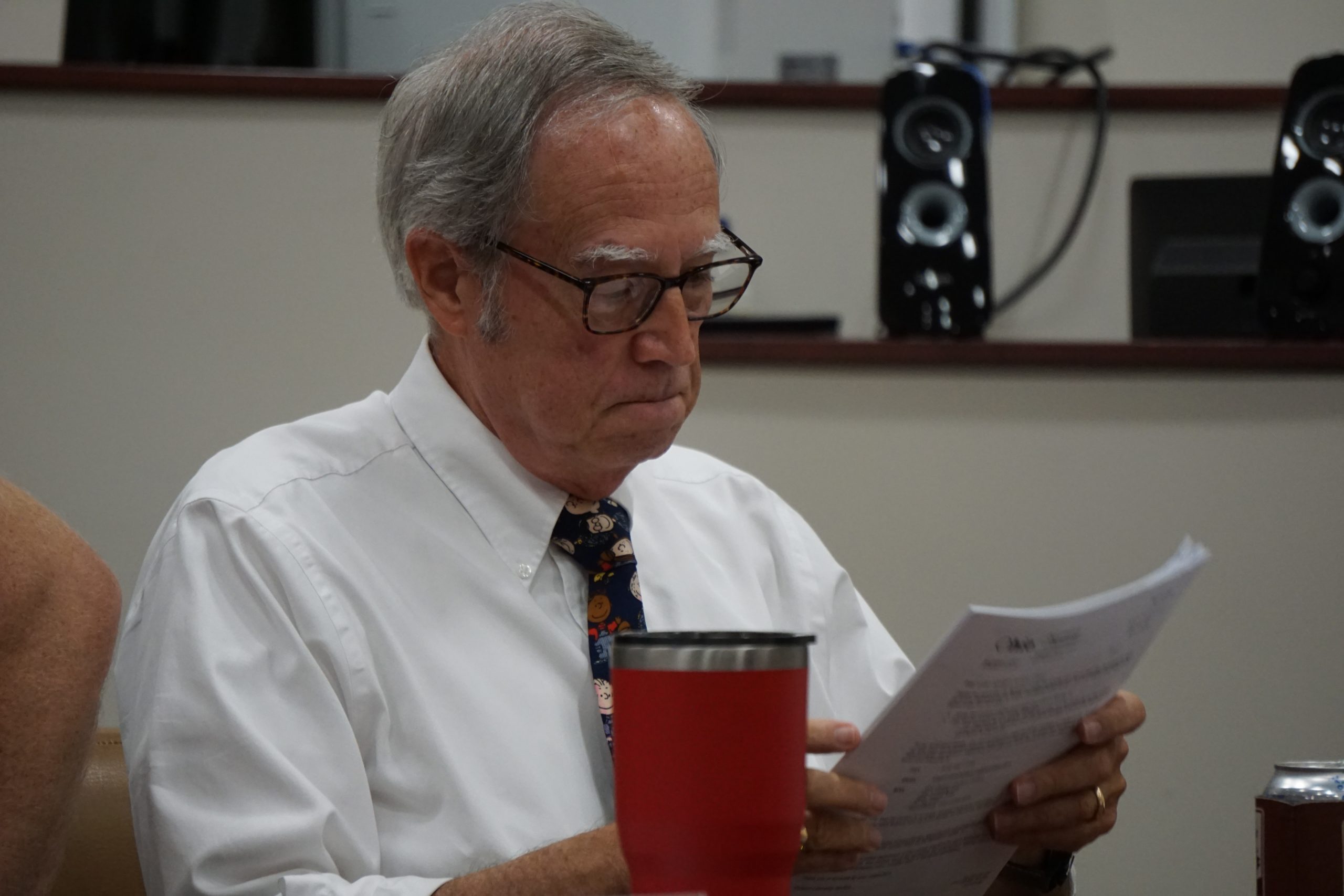 Man in white shirt, tie, and wearing glasses reads from a piece of paper in his hands.