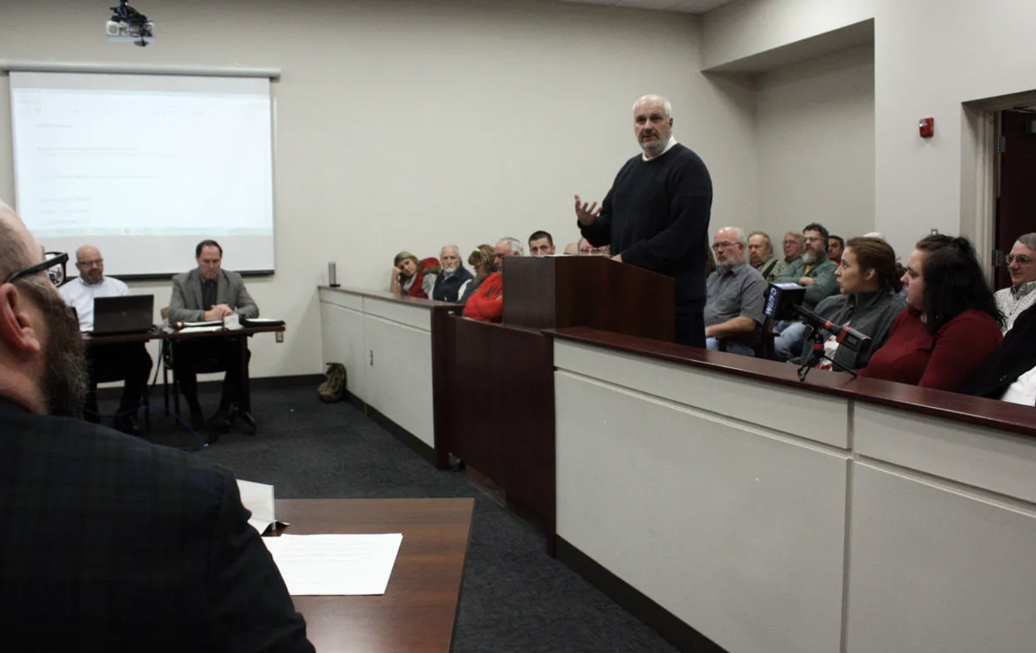 Man in black sweater speaking at podium during city council meeting.