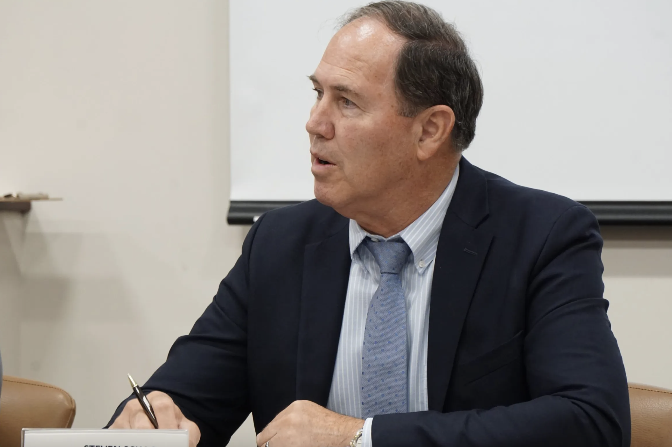 Man in suit sits at a desk and holds a pen in his right hand.