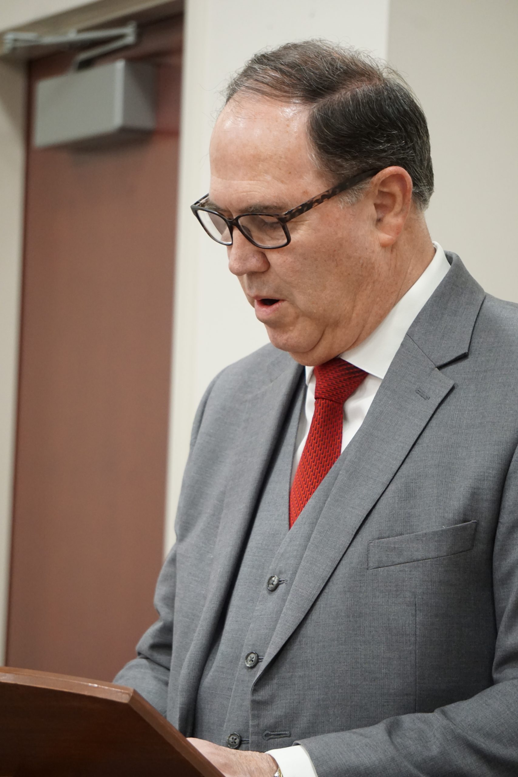 Man, wearing suit and glasses, giving speech at podium.